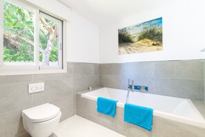 A bathroom with a bathtub, a toilet, and a window with a view of greenery.