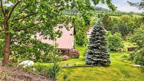 A rural landscape with a large tree, a building with a red roof, and a coniferous tree in the foreground.