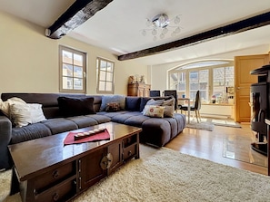 A living room with a dark-colored sofa, a wooden coffee table, and a fireplace. There are two windows and a door leading to an outdoor area.