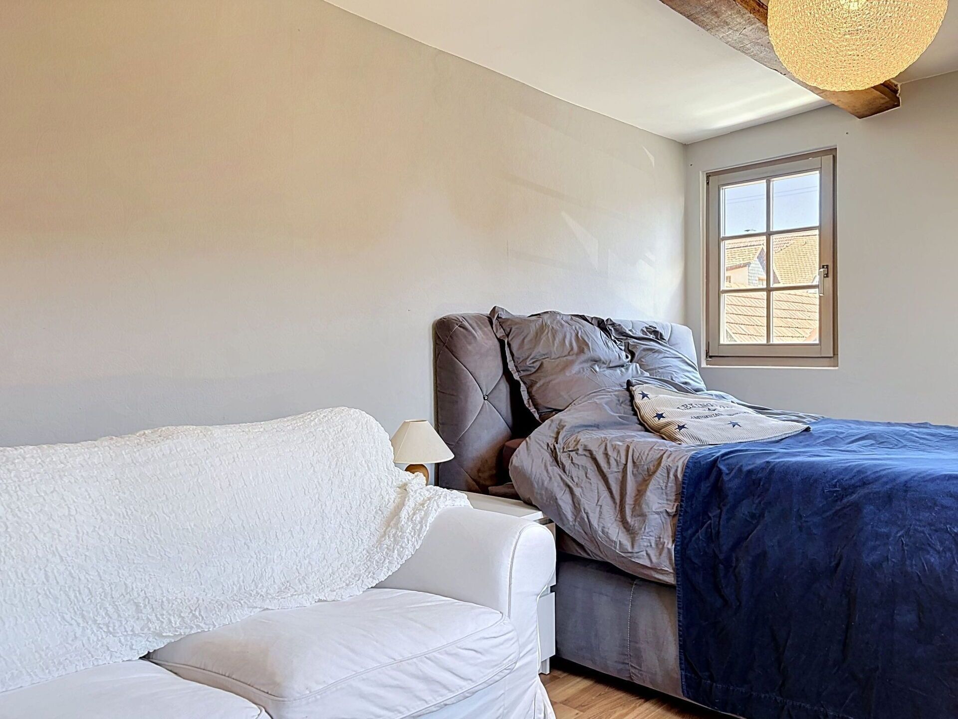A bedroom with a bed, a white sofa, a lamp, a window with a view, and a textured ceiling.