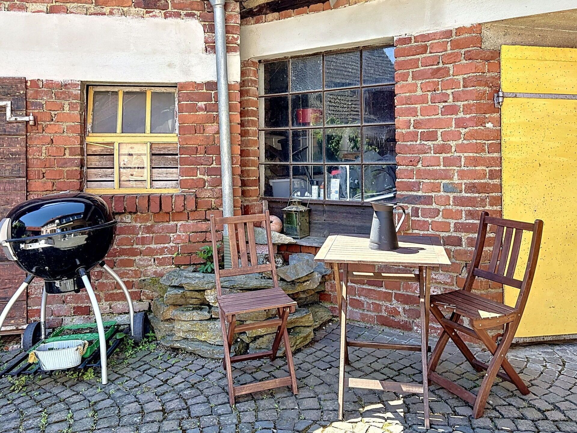 A brick building with a yellow door, a window with a wooden shutter, a barbecue grill, and a table with chairs outside.