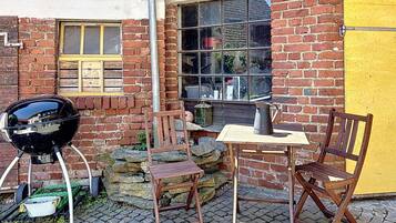 A brick building with a yellow door, a window with a wooden shutter, a barbecue grill, and a table with chairs outside.