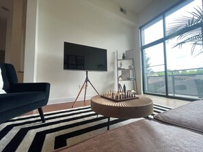 Ein modernes Wohnzimmer mit einem großen Fernseher auf einem Dreibein-Ständer, einem Schachbrett, einem runden Hocker und einem großen Fenster mit Blick auf Grünflächen.