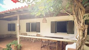 Une terrasse couverte avec une table et des chaises, un arbre et un bâtiment doté de fenêtres.
