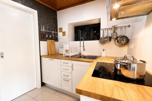 Een kleine keuken met witte kastjes, een houten aanrecht, een fornuis, een spoelbak en hangend keukengerei.