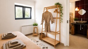 Une pièce comprenant un lit, un étendoir en bois, un radiateur, une fenêtre et une salle de bain avec un lavabo et un miroir.