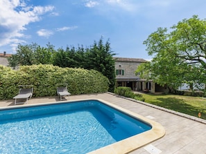 Ein rechteckiger Pool mit Steinbord, zwei Liegestühle, ein Steinhaus mit Ziegeldach und ein gepflegter Heckenstreifen.