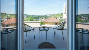 Un balcone con sedie e tavolo, vista su una cittadina con case e una statua, e cielo sereno.