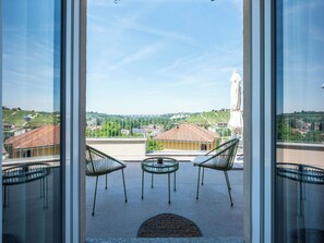 Un balcone con sedie e tavolo, vista su una cittadina con case e una statua, e cielo sereno.