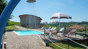 Area piscina con doccetta, poltroncine, ombrelloni e un edificio sullo sfondo.