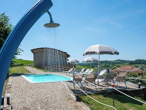 Area piscina con doccetta, poltroncine, ombrelloni e un edificio sullo sfondo.