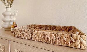 Ein geflochtener Korb auf einer Holztischplatte mit einer Vase und einem Schilf-Diffusor im Hintergrund.