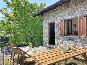 Ein Steinhaus mit Holztisch und -stühlen, einem Grill und Blick auf eine baumbewachsene Gegend.