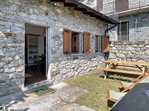 Ein Steinhaus mit Holztür, Fenstern mit Fensterläden, einer Terrasse mit Tisch und Stühlen und einem kleinen Garten.