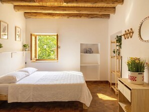 Ein Schlafzimmer mit Bett, Fenster, Holzbalkendecke, Ablage mit Handtüchern, Spiegel und Pflanze.