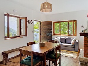 Ein gemütliches Wohnzimmer mit einem Holzesstisch, einer Couch und großen Fenstern mit Blick ins Grüne.