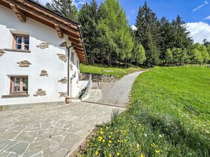 Ein traditionelles Chalet mit Steinwänden, einer Holztreppe und einem Kiesweg, der zu einer Wiese mit Wildblumen führt.