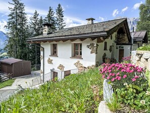 Ein rustikales Chalet mit Steinmauern, Holzbalken und einem Steinkamin. Es verfügt über einen kleinen Balkon, eine Holzterrasse und einen Garten mit rosa Blumen.