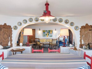 Ein gemütliches Zimmer mit einem Bett, zwei Sesseln, einer Couch und einem Esstisch. An der Wand befinden sich dekorative Teller und eine rote Lampe hängt von der Decke.