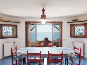 Ein Essbereich mit Tisch und Stühlen, einer Hängelampe und Blick auf das Meer durch eine Tür.
