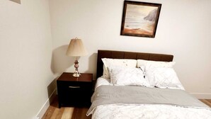 Un letto ben rifatto con un copriletto bianco e cuscini, un comodino con una lampada e un quadro appeso al muro.