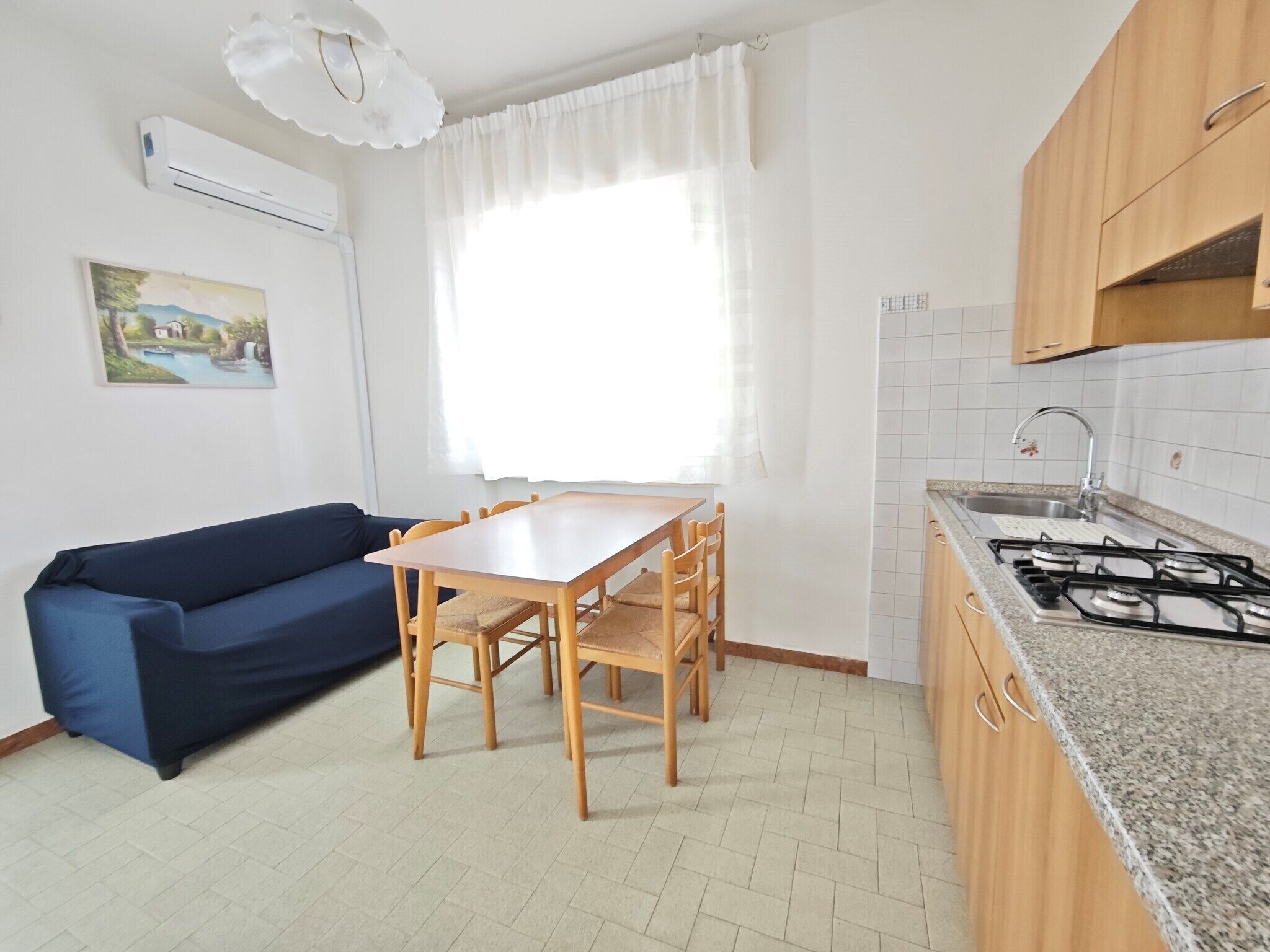 A compact kitchen with a gas stove, a dining table with chairs, a blue sofa, and a window with sheer curtains.
