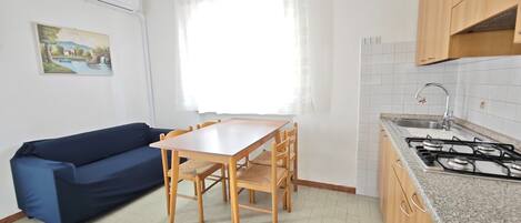 A compact kitchen with a gas stove, a dining table with chairs, a blue sofa, and a window with sheer curtains.