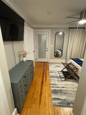 Quarto com piso de madeira, penteadeira cinza, cama com cobertor azul, ventilador de teto e espelho na parede.