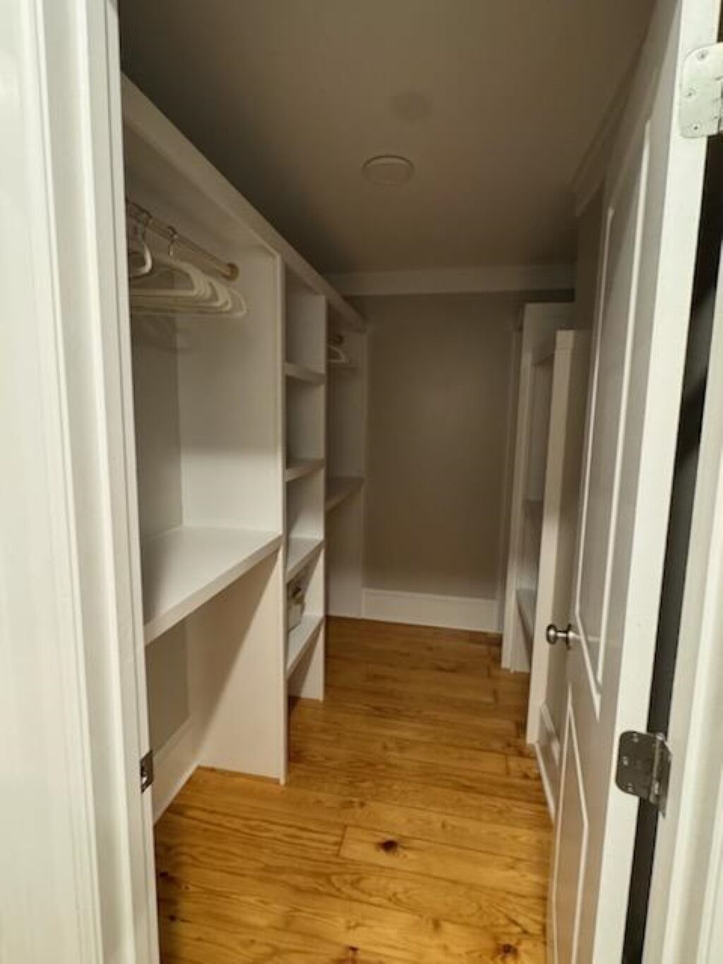 A narrow hallway with white closet doors and shelves, wooden flooring, and a white wall.