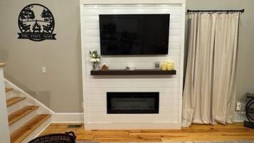 A living room with a flat-screen TV mounted on a white mantel, a fireplace below, and a staircase to the left.