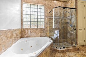 Un baño con bañera, cabina de ducha de vidrio y ventana con celosías decorativas.