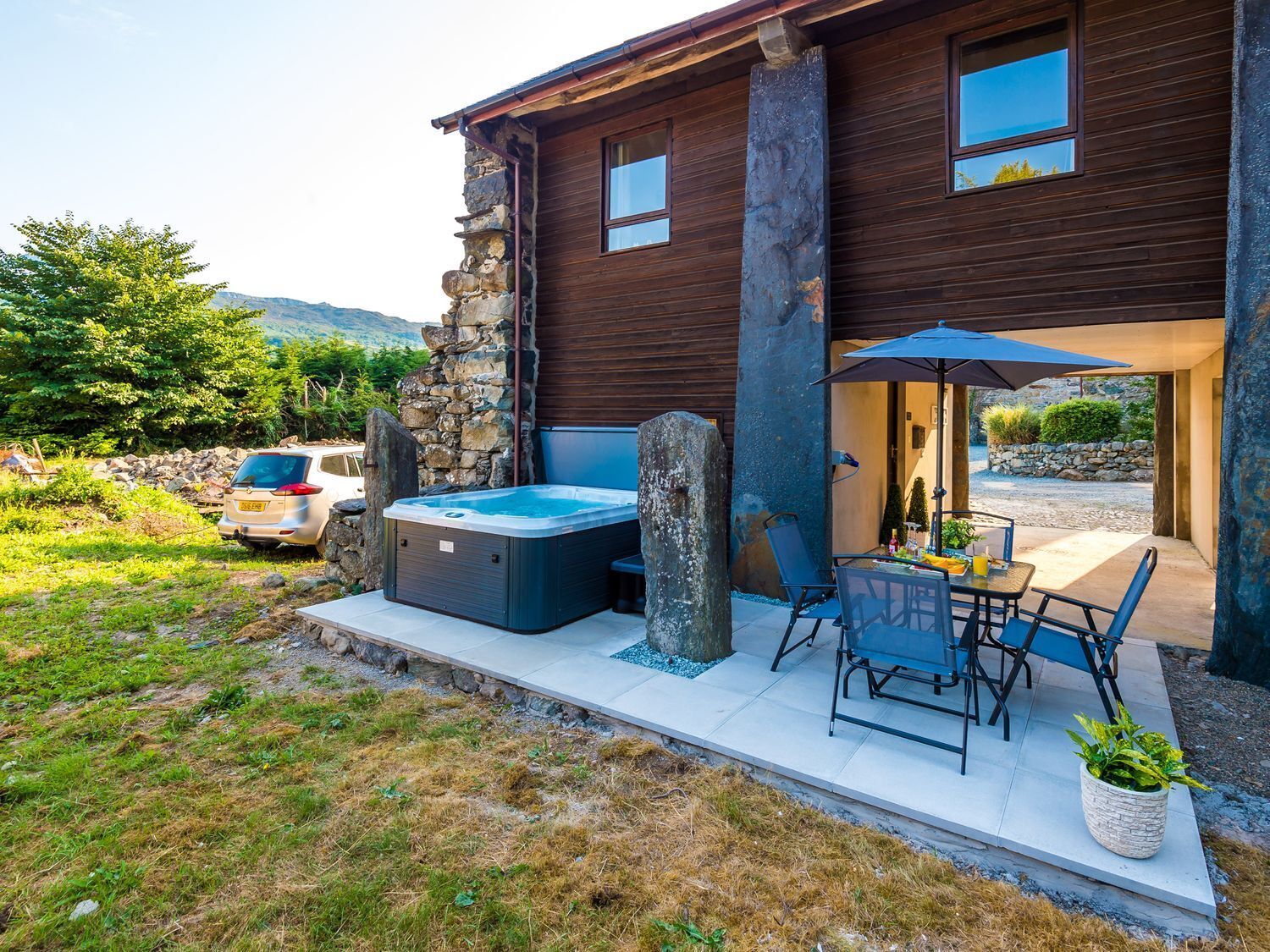 Una casa moderna con un muro de piedra, un jacuzzi, mobiliario exterior y un coche aparcado cerca.