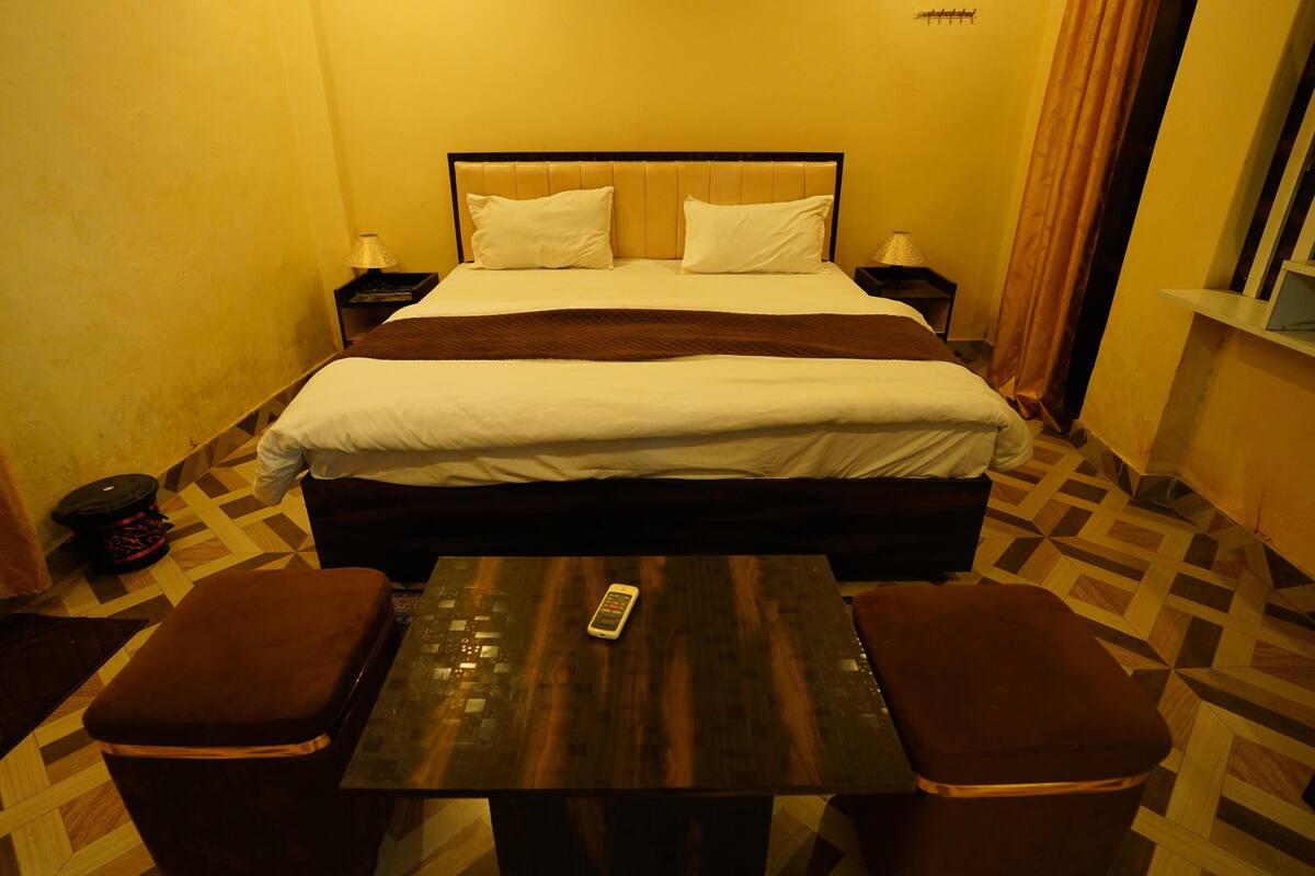 Habitación de hotel con una cama, mesitas de noche, una mesa con un control remoto y dos zonas de estar con cojines.
