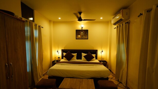 A hotel room with a bed, bedside lamps, a ceiling fan, an air conditioning unit, and a small wooden table. - The Grand Ganga Paying Guest (VARANASI)