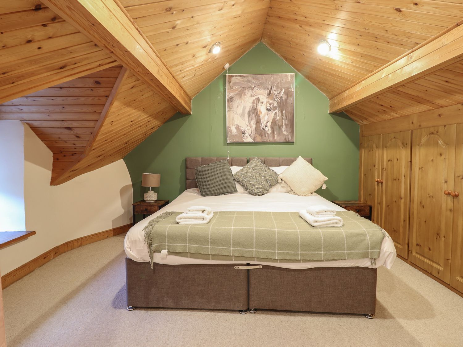 Un dormitorio con techo inclinado, una cama con edredón verde a cuadros, varias almohadas, una mesita de noche con lámpara y un armario de madera.