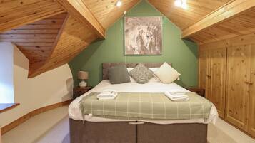 Un dormitorio con techo inclinado, una cama con edredón verde a cuadros, varias almohadas, una mesita de noche con lámpara y un armario de madera.