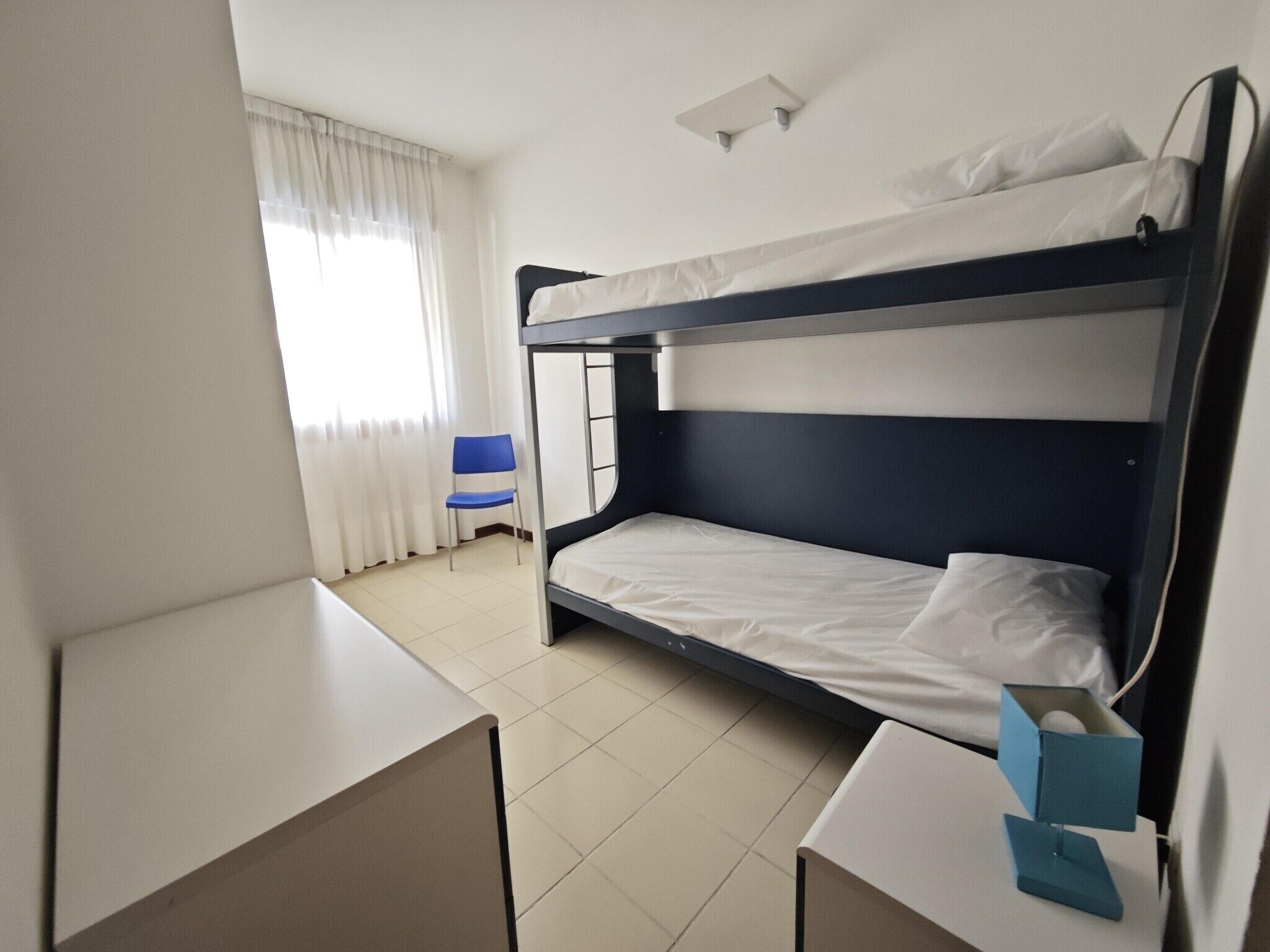 A bunk bed room with a desk and chair, a window with curtains, and a ceiling light fixture.