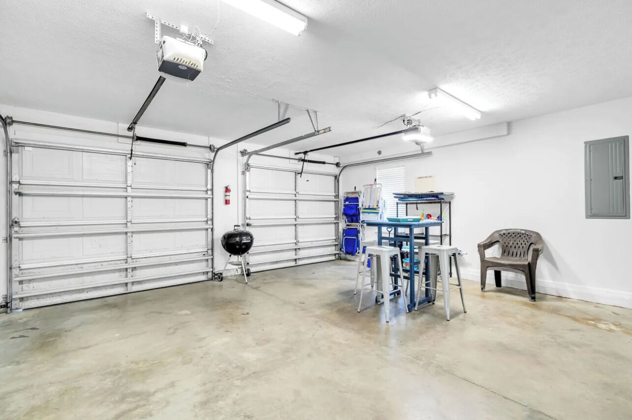 A garage with two white garage doors, a ceiling-mounted projector, a fire extinguisher, a barbecue grill, and a workbench with tools.