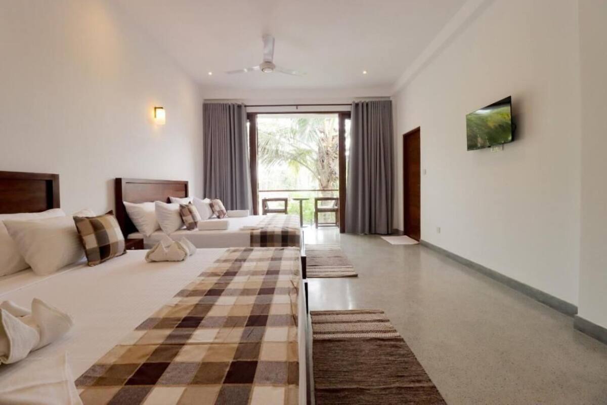 Habitación de hotel con dos camas, ventilador de techo, televisor y balcón con cortinas.