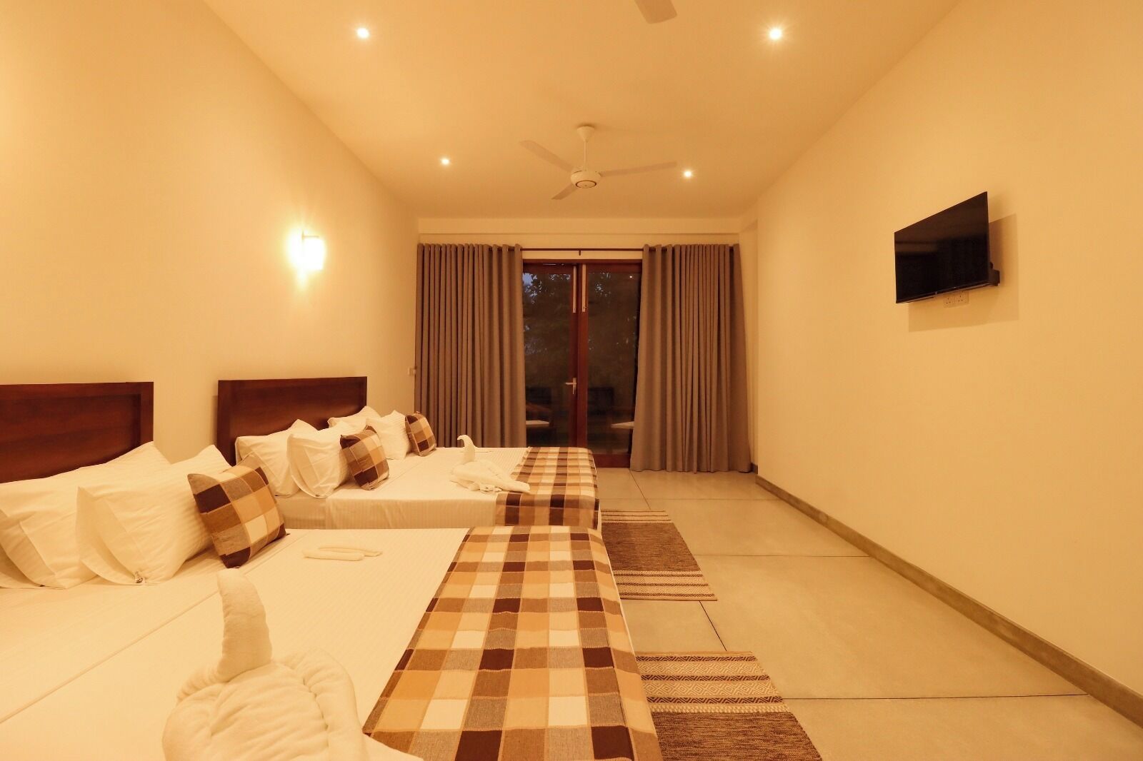 A hotel room with two beds, a TV, a ceiling fan, and a view of the outdoors through the window.