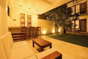 A cozy indoor seating area with a sofa, chairs, and a coffee table, a small bar area with bottles, and a well-lit outdoor space with a tree and a building in the background.