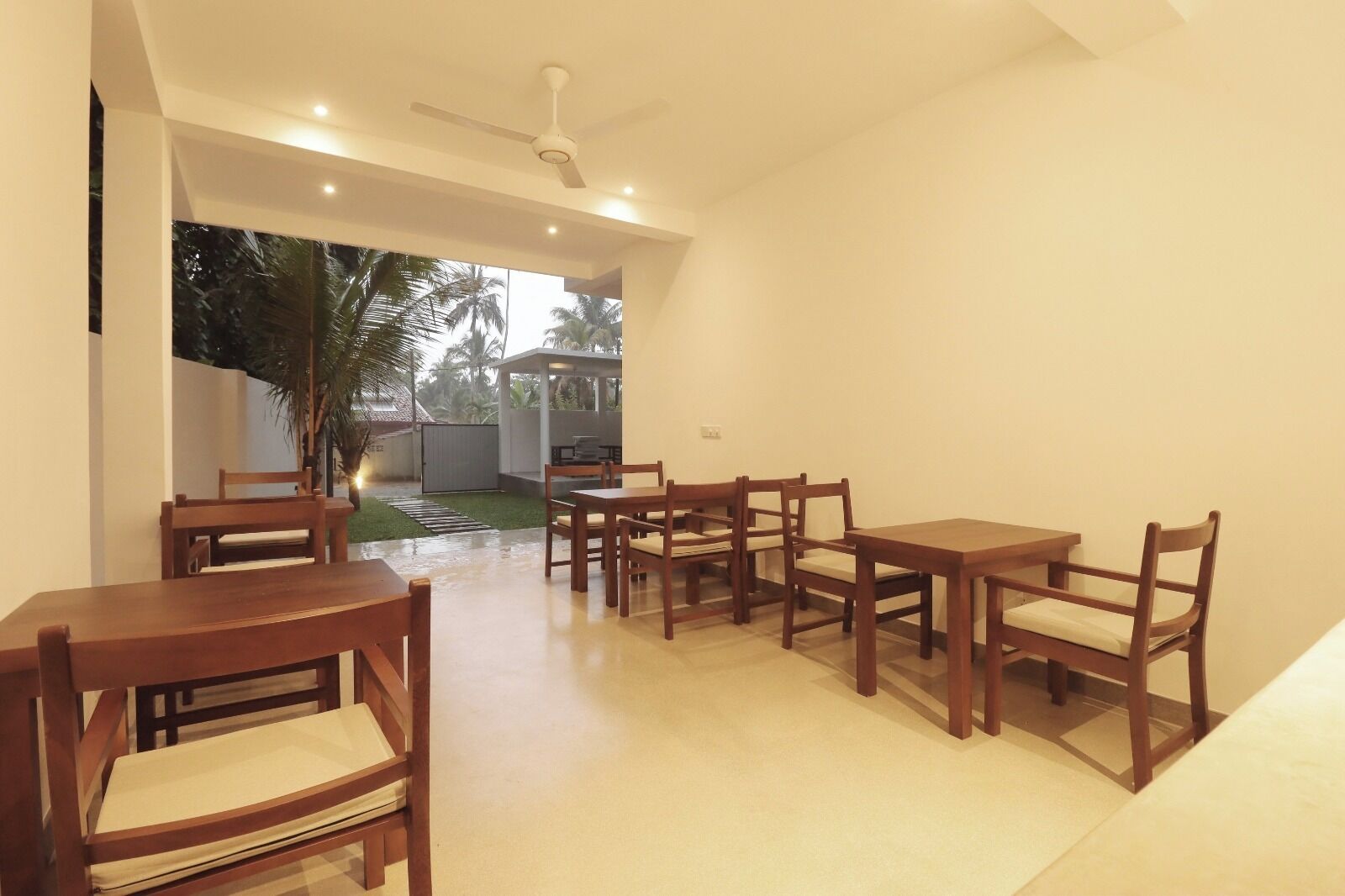 A dining area with wooden tables and chairs, a ceiling fan, and a view of a garden with palm trees.