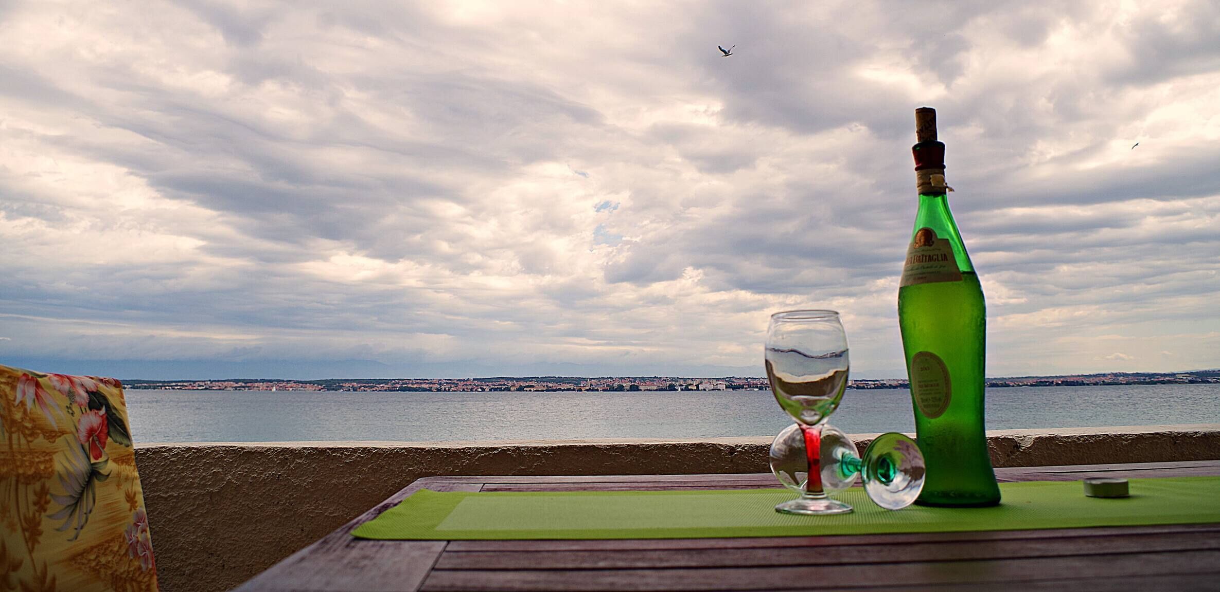 A table with a bottle and a glass, overlooking a body of water and a distant cityscape.