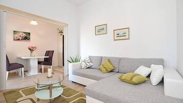 A modern living room with a grey sofa, a glass coffee table, a dining table with chairs, and a small plant.