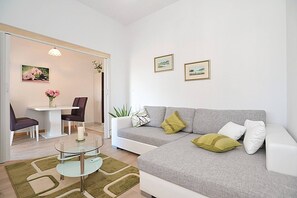 Ein modernes Wohnzimmer mit einer grauen Couch, einem Glastisch, einem Esstisch mit Stühlen und einer kleinen Pflanze.