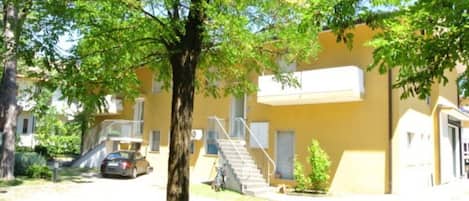 Un bâtiment jaune avec un escalier menant à une porte, entouré d’arbres et avec une voiture garée à proximité.