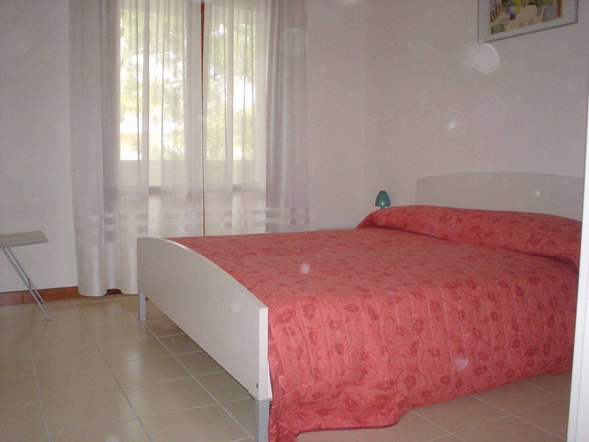 A bedroom with a bed, a nightstand, a lamp, and a window with sheer curtains.