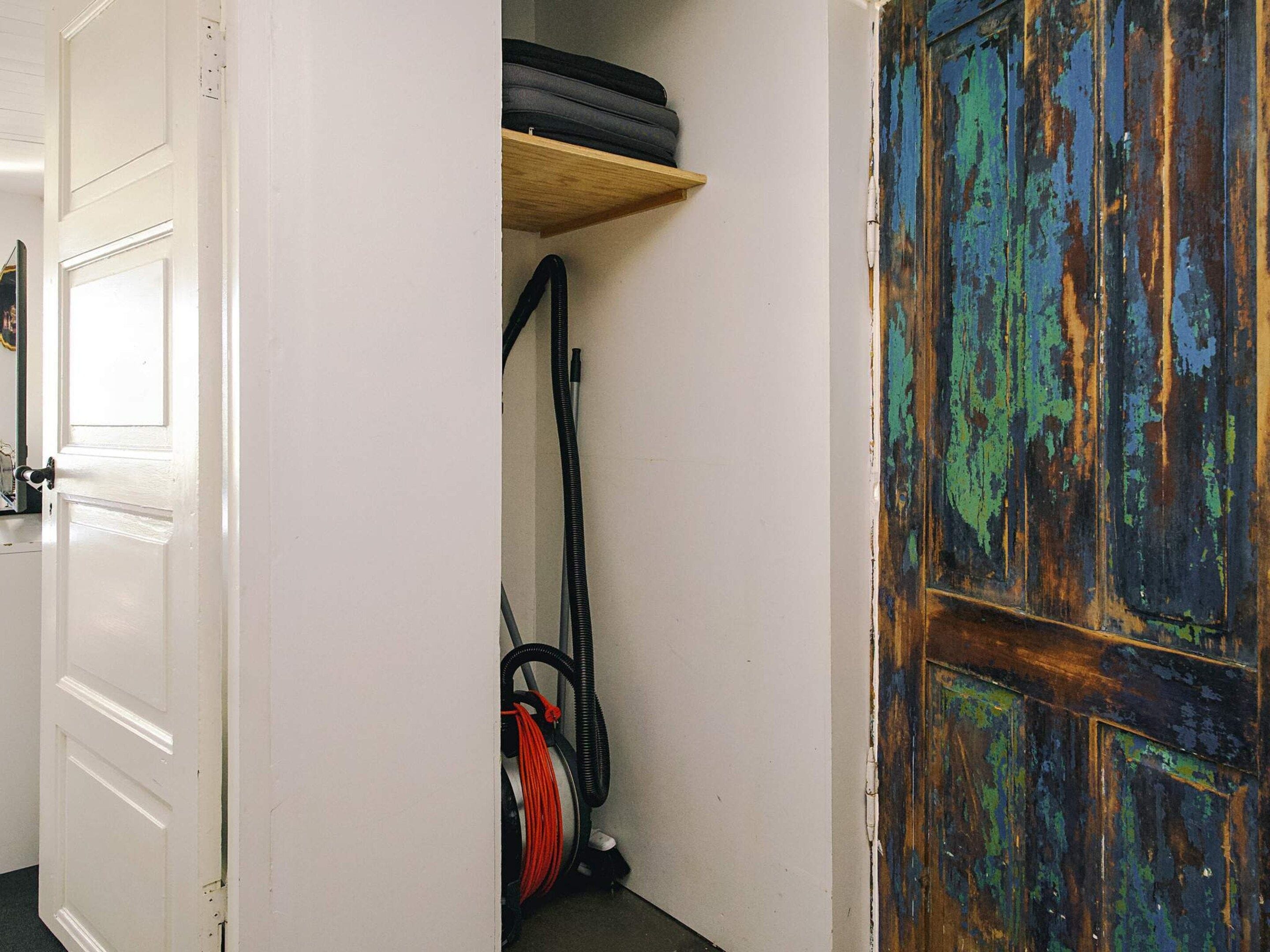 Ein schmaler Flur mit einer weißen Tür links und einer rustikalen Holztür rechts. Eine Staubsaugerin ist in der Ecke sichtbar.