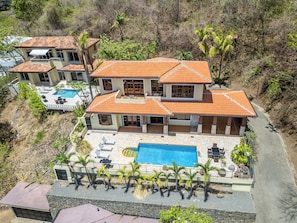 Ein großes, zweistöckiges Haus mit Swimmingpool, mehreren Balkonen und einem angelegten Garten mit Palmen.