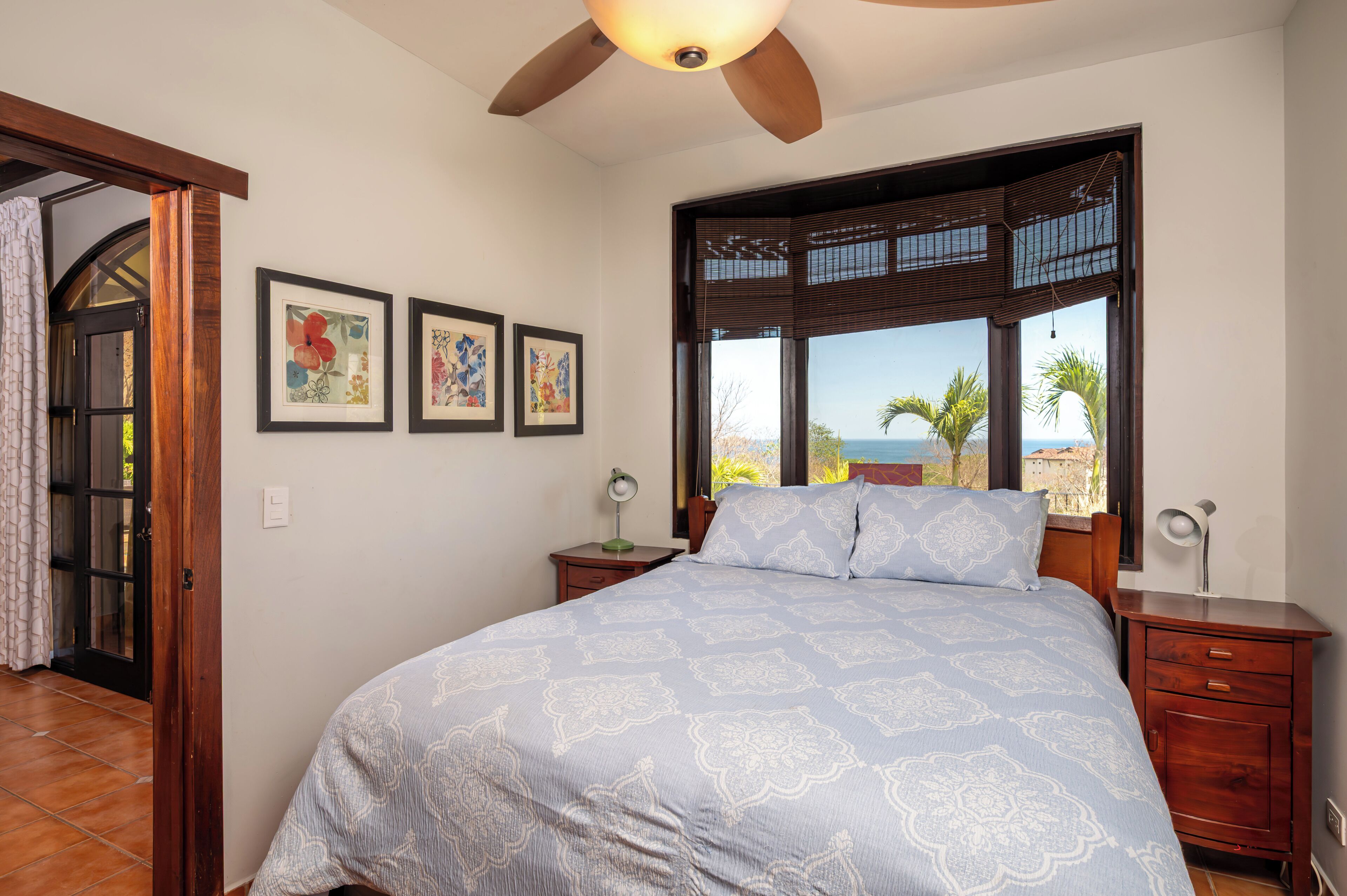 A bedroom with a bed, a nightstand, a desk, a ceiling fan, a window with a view, and framed artwork on the wall.